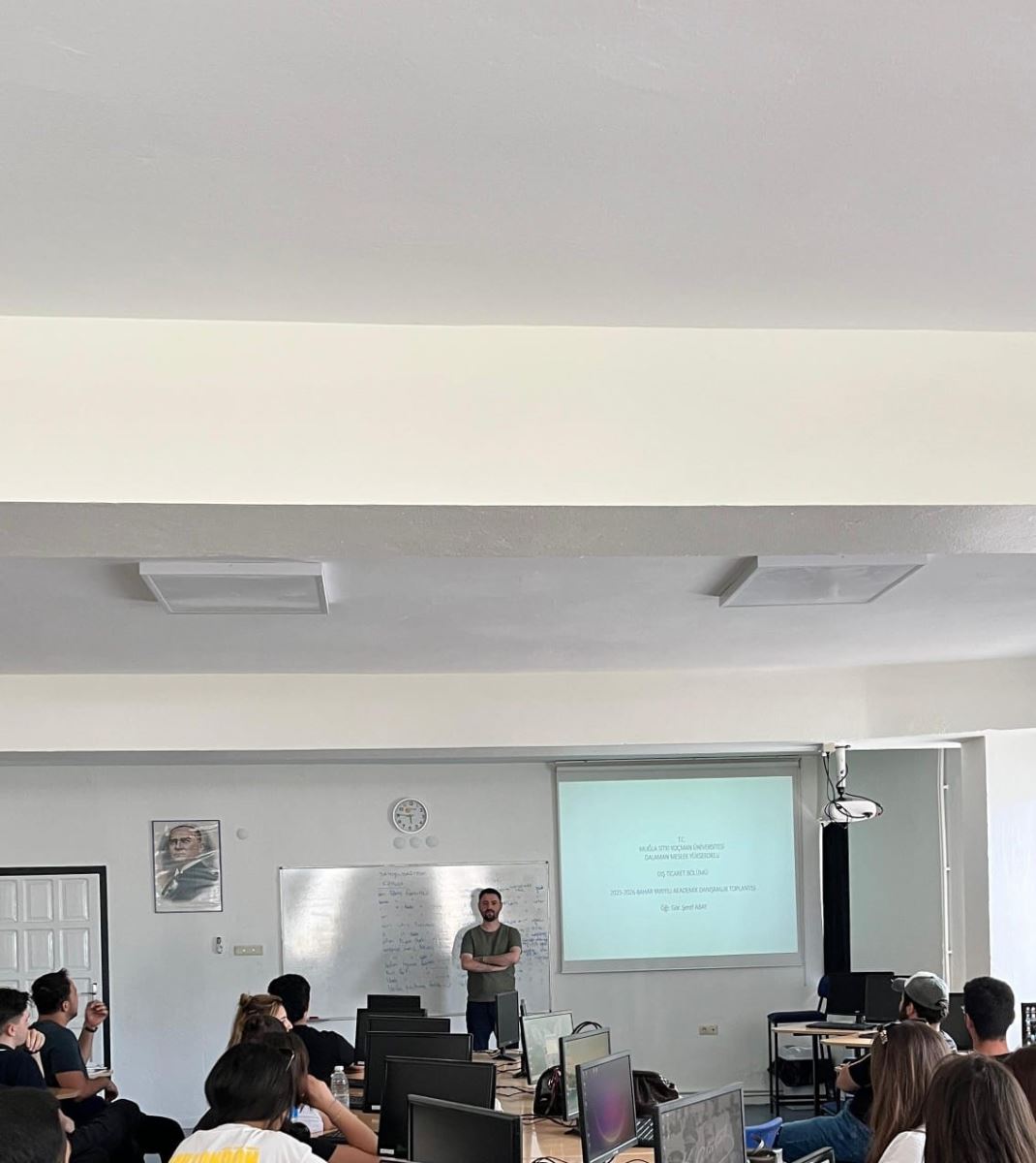 In a bright computer laboratory, approximately twenty students are seated at semicircular desks. On the desks are black computer monitors, keyboards, and mice. Near the camera, an upside-down computer monitor rests on a desk.  At the front of the classroom, in front of a whiteboard and a projection screen, an instructor is giving a lecture. A formal meeting-style presentation is projected onto the screen.  The students are attentively looking forward; some are resting their elbows on the desks, while others are slightly turned toward the instructor.  The classroom has white walls and a high ceiling. On the front wall, there is a clock and a framed portrait of Mustafa Kemal Atatürk. The overall atmosphere is calm and focused on learning.